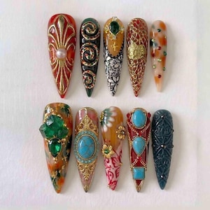 May include: An assortment of ten long, pointed artificial nails. Each nail features intricate designs, including gold filigree, gemstones, and detailed patterns in various colors like red, green, and turquoise. The nails are arranged on a white surface.