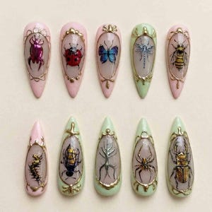May include: A set of ten almond-shaped artificial nails. Each nail features a different insect design, including a beetle, ladybug, butterfly, dragonfly, and bee, set against a pink or green background. The designs are framed with gold filigree.