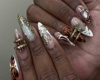 May include: A set of long, pointed fingernails with intricate designs. The nails feature a variety of colors, including white, brown, and gold, with ornate gold embellishments and floral patterns. Some nails have a clear base with detailed designs.