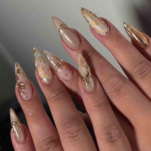 May include: A close-up of a hand with long, stiletto-shaped nails. The nails are painted in a nude color and adorned with gold accents. Some nails feature gold embellishments, while others have a marbled effect with gold trim. The overall design is elegant and glamorous.