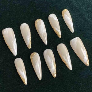 May include: Set of ten artificial nails with a long, pointed shape. Each nail features a creamy white base with a marbled effect and gold metallic accents along the edges. The nails are arranged on a dark green surface.