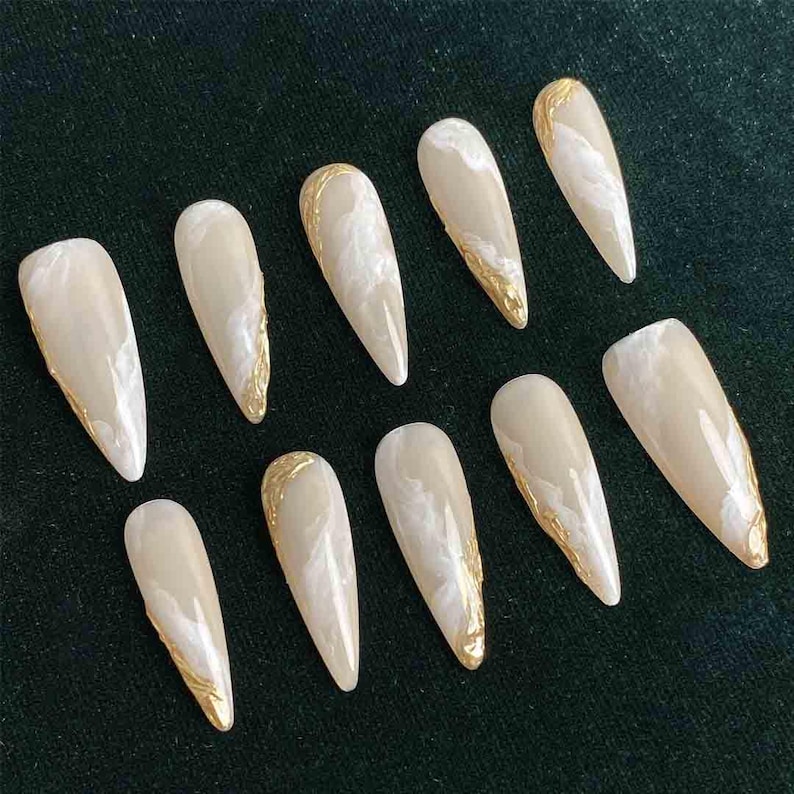 May include: Set of ten artificial nails with a long, pointed shape. Each nail features a creamy white base with a marbled effect and gold metallic accents along the edges. The nails are arranged on a dark green surface.