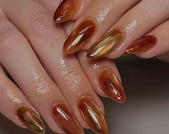Könnte beinhalten: Nahaufnahme einer Hand mit langen, mandelförmigen Nägeln. Die Nägel sind mit einem glänzenden, warmbraunen Lack bemalt, der einen Cat-Eye-Effekt hat und ein schimmerndes, metallisches Aussehen erzeugt. Die Nägel reflektieren das Licht und zeigen die Tiefe und Dimension der Farbe.