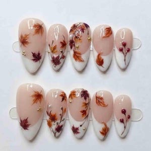 May include: A set of ten almond-shaped press-on nails featuring a nude base with white tips. The design includes hand-painted autumn leaves in shades of orange, red, and brown, accented with small gold metallic beads and delicate berry sprigs.