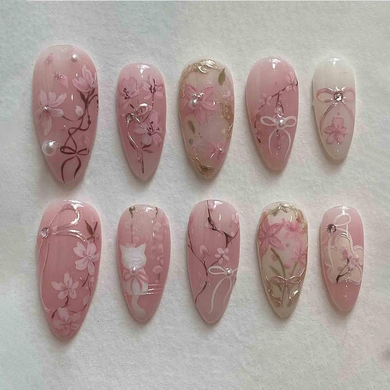 May include: A set of ten almond-shaped artificial nails. Each nail is painted with a light pink base and features floral designs, bows, and silver accents. Some nails have a white cat design. The overall aesthetic is delicate and feminine.