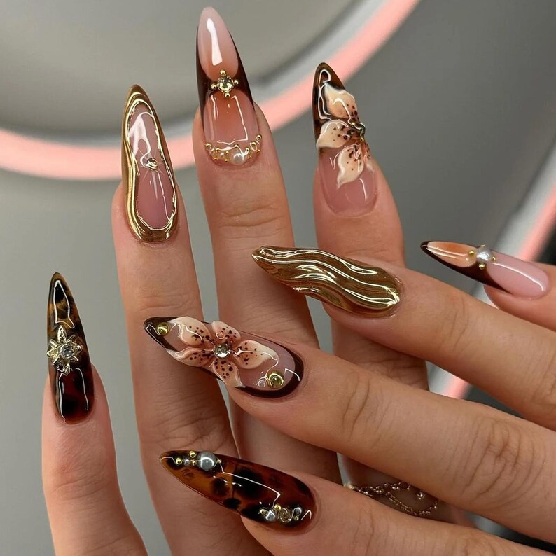 May include: A close-up of a hand with long, almond-shaped acrylic nails. The nails feature a variety of designs, including floral accents, gold embellishments, and a tortoiseshell pattern. The color palette includes brown, gold, and pink tones.