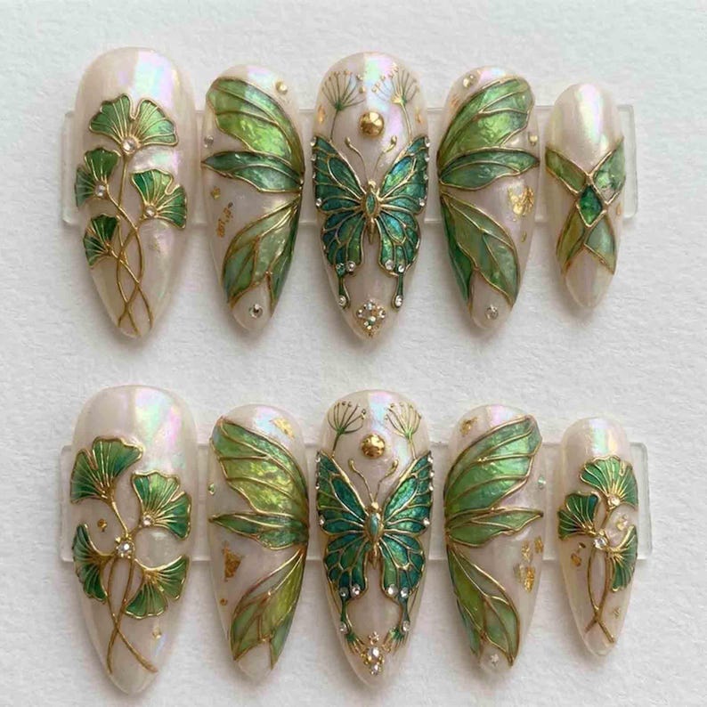 Puede incluir: Un juego de diez u&ntilde;as postizas en forma de almendra con una base blanca iridiscente. Los dise&ntilde;os incluyen alas de mariposa verdes y doradas, motivos de hojas de ginkgo y peque&ntilde;os detalles dorados. Cada u&ntilde;a tiene l&iacute;neas doradas met&aacute;licas.