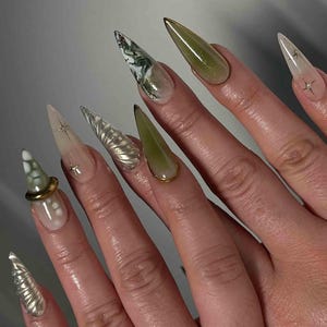 May include: A hand displaying long, pointed fingernails with various designs. Some nails are clear with star accents, others feature green and white marble patterns, and some are a solid olive green. One nail has a gold ring accent.