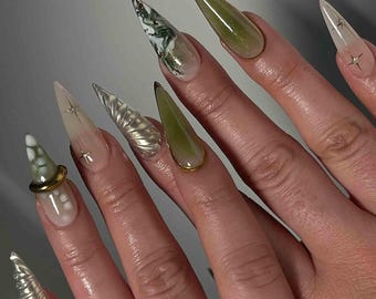 May include: A hand displaying long, pointed fingernails with various designs. Some nails are clear with star accents, others feature green and white marble patterns, and some are a solid olive green. One nail has a gold ring accent.