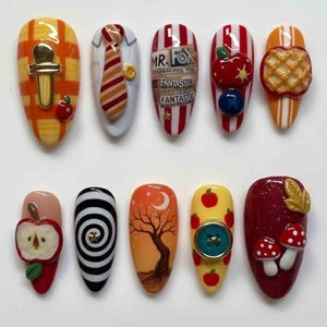 Op de afbeelding: Een set van tien kunstnagels met diverse ontwerpen. De ontwerpen omvatten een gouden sluiting, een stropdas, de tekst "Mr. Fox", appels, een wafel, een spiraal, een boom, een knoop en paddenstoelen.