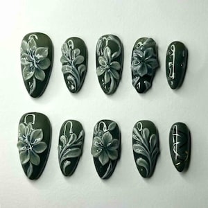 May include: Set of ten olive green press-on nails with 3D floral designs. Each nail features a different design, including flowers and leaf patterns in shades of gray and white. The nails are almond-shaped.
