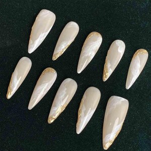 May include: A set of ten almond-shaped false nails. Each nail is a light beige color with a marbled white design and gold foil accents along the edges. The nails are arranged on a dark green surface.