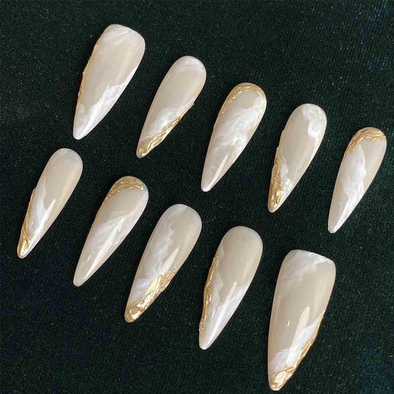 May include: A set of ten almond-shaped false nails. Each nail is a light beige color with a marbled white design and gold foil accents along the edges. The nails are arranged on a dark green surface.