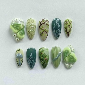 May include: A set of ten almond-shaped artificial nails with nature-inspired designs. The nails feature various shades of green, with detailed artwork including a butterfly, mushrooms, branches, and leaves. Some nails have 3D elements.