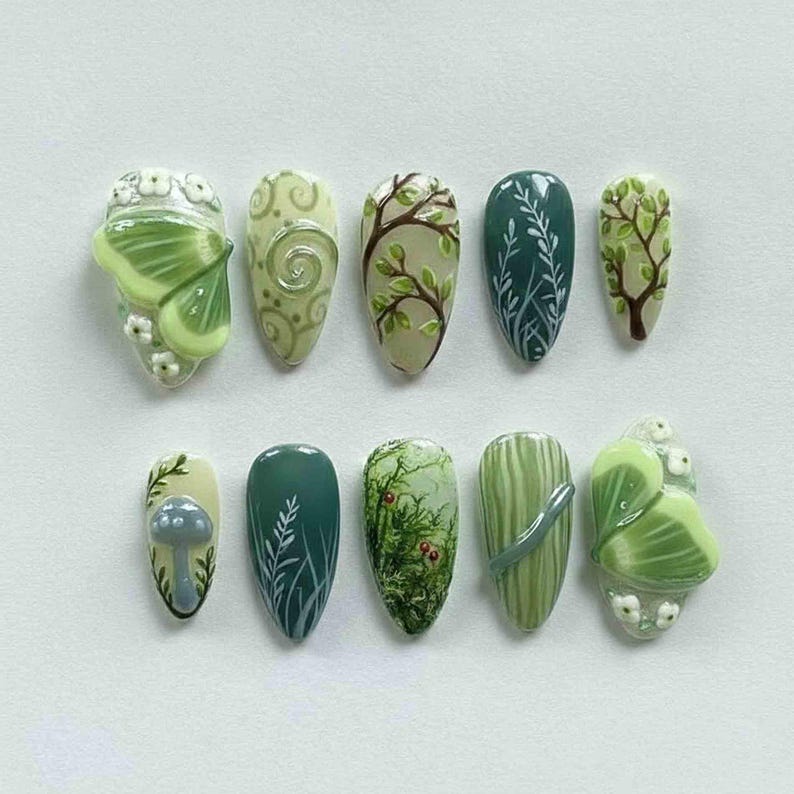 May include: A set of ten almond-shaped artificial nails with nature-inspired designs. The nails feature various shades of green, with detailed artwork including a butterfly, mushrooms, branches, and leaves. Some nails have 3D elements.