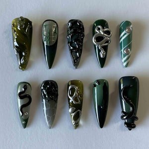 May include: A collection of ten long, pointed artificial nails. Each nail features a unique design, including swirling patterns, geometric shapes, and metallic accents. Colors range from olive green to deep forest green, with silver and black embellishments.
