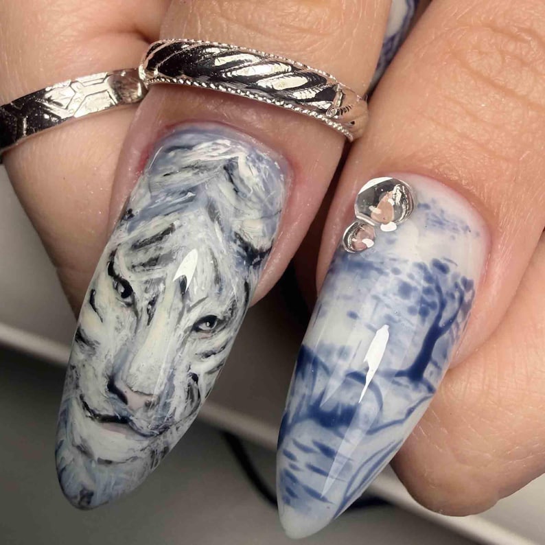 May include: Almond-shaped fingernails featuring detailed nail art. One nail displays a white tiger face in shades of white, gray, and black. The adjacent nail features a blue and white landscape scene with a tree and two small clear rhinestones.