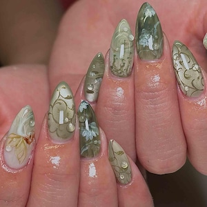 May include: A close-up of a hand with long, almond-shaped nails. The nails are painted with a green and white floral design, with water droplets on the surface. The design includes swirls and leaf patterns, creating an elegant and artistic look.