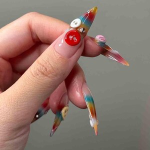 Puede incluir: U&ntilde;as acr&iacute;licas largas y puntiagudas con un dise&ntilde;o de degradado arco&iacute;ris. Cada u&ntilde;a est&aacute; adornada con botones decorativos de varios colores y tama&ntilde;os, incluyendo rojo, blanco y rosa. Algunas u&ntilde;as presentan peque&ntilde;os diamantes de imitaci&oacute;n. Las u&ntilde;as son una declaraci&oacute;n de moda &uacute;nica y art&iacute;stica.