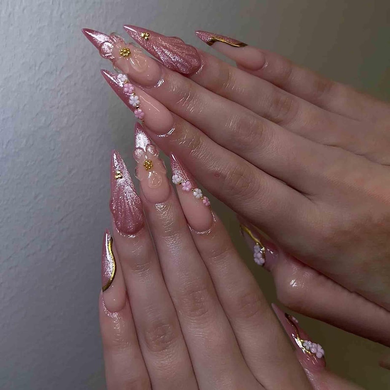 May include: A set of long, pointed acrylic nails. The nails are painted in a light pink hue with glitter accents. Some nails feature gold embellishments and small, white and pink floral designs.