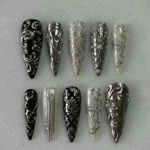 May include: A collection of long, pointed nail covers with elaborate designs. The colours are black, silver, and white. Some have detailed scrollwork, others pearl accents. The nail covers are arranged in two rows, showcasing different styles.