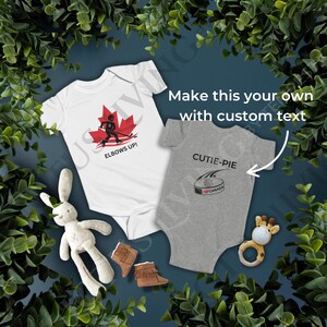 Può includere: Due body per neonati bianchi e grigi con disegni a tema hockey. Un body presenta un giocatore di hockey con una foglia d'acero rossa e il testo "Elbows Up!" L'altro body presenta un disco di hockey con il testo "Cutie-Pie" e una bandiera canadese.