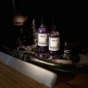 May include: Two small glass bottles with purple liquid and black labels. The labels read "Coven 13" and "Morning and Body Oil" and "Dark Moon Trading Co."