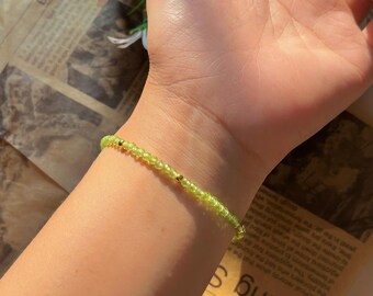 May include: A delicate bracelet featuring small, light green beads strung together with gold-colored accents. The bracelet is worn on a wrist, showcasing its subtle elegance. The background includes a newspaper clipping and white flowers.