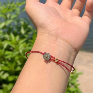 May include: A red string bracelet with a flower-shaped jade charm and gold accents. The bracelet is tied with a sliding knot for adjustable sizing. The jade charm is a light green color.