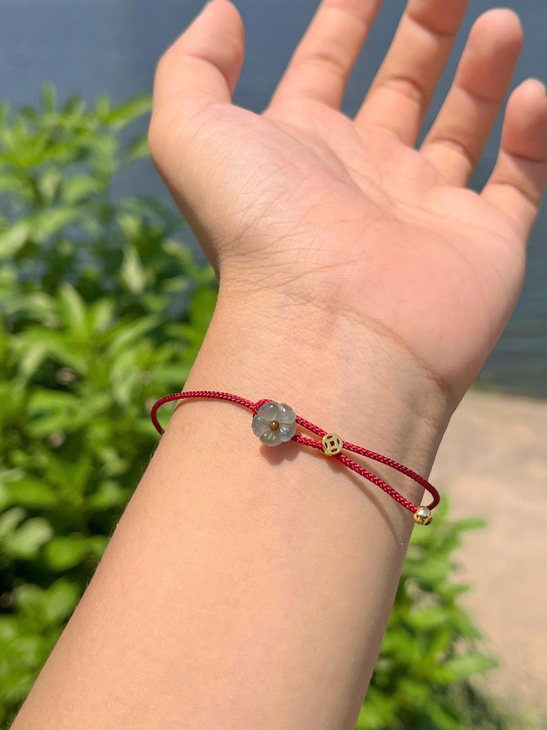May include: A red string bracelet with a flower-shaped jade charm and gold accents. The bracelet is tied with a sliding knot for adjustable sizing. The jade charm is a light green color.