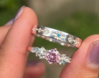 May include: Two silver rings. One ring features a row of stars and arrows. The other ring has a large pink gemstone surrounded by smaller white and blue gemstones. The rings are held up by a person's hand.