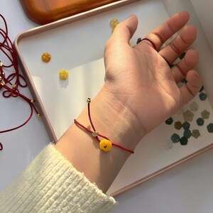 May include: A red string bracelet with a small, round, yellow charm is worn on a wrist. The bracelet has a delicate design with a gold-colored bead. The background includes a white surface and a wooden tray.