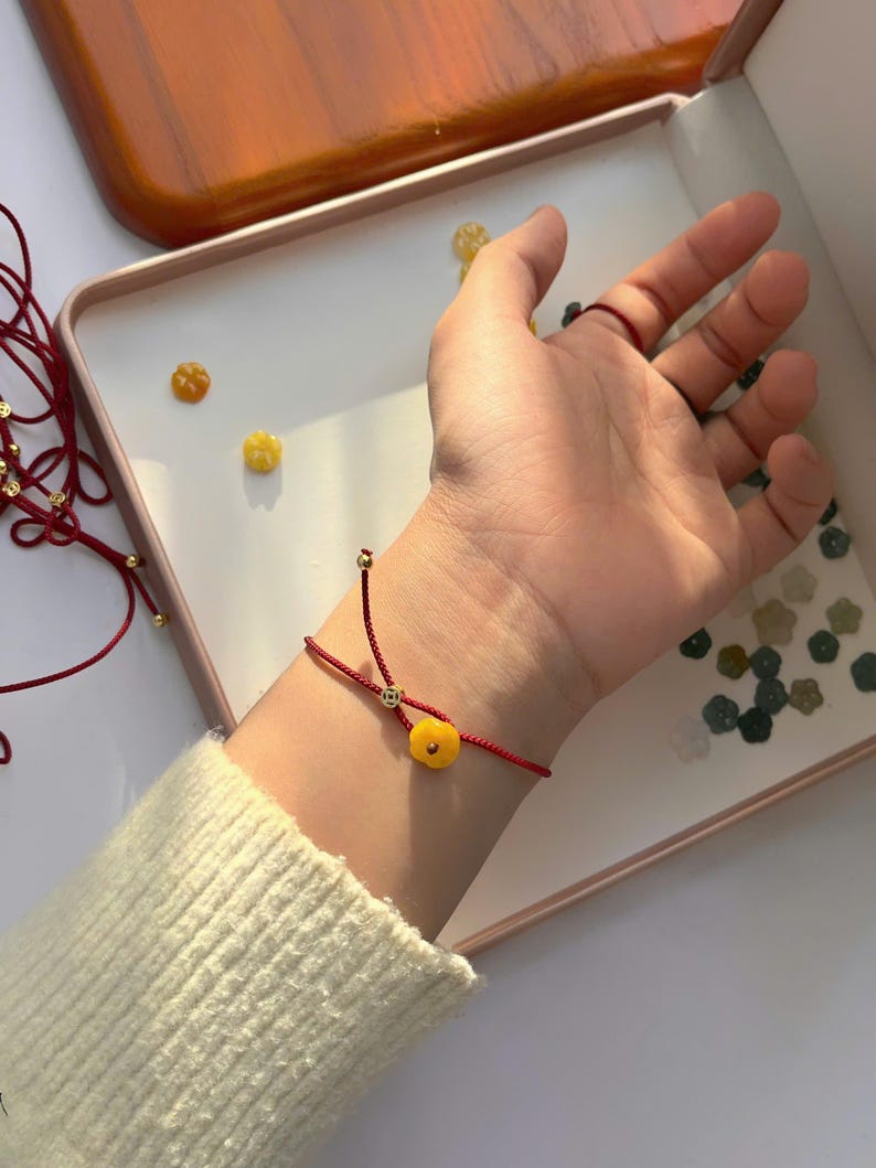 May include: A red string bracelet with a small, round, yellow charm is worn on a wrist. The bracelet has a delicate design with a gold-colored bead. The background includes a white surface and a wooden tray.