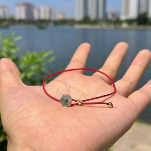 May include: A red cord bracelet featuring a light green flower-shaped charm and two small gold beads. The bracelet is displayed against a blurred background of a lake and buildings.