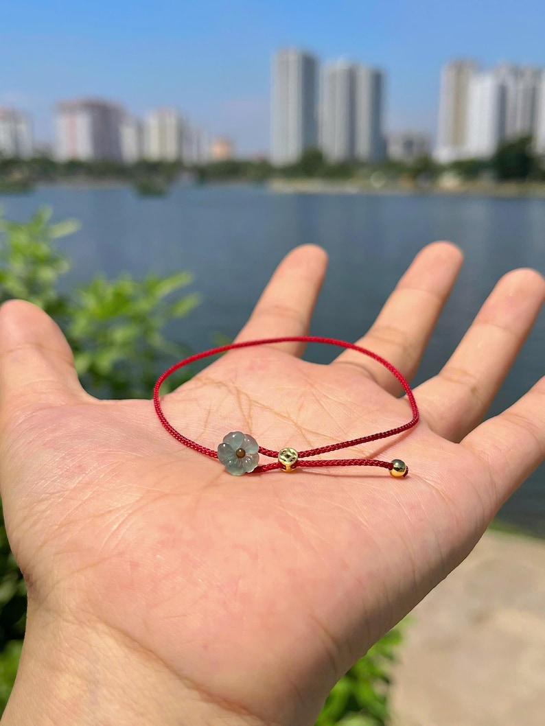 May include: A red cord bracelet featuring a light green flower-shaped charm and two small gold beads. The bracelet is displayed against a blurred background of a lake and buildings.
