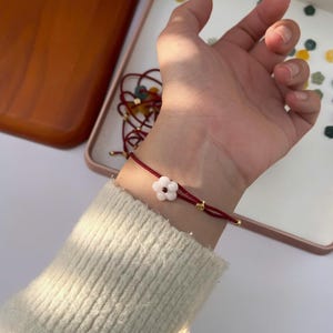 May include: A red string bracelet with a white flower charm. The bracelet features gold-colored accents and is worn on a wrist. The background includes a white surface and a tray with small, colorful decorative elements.