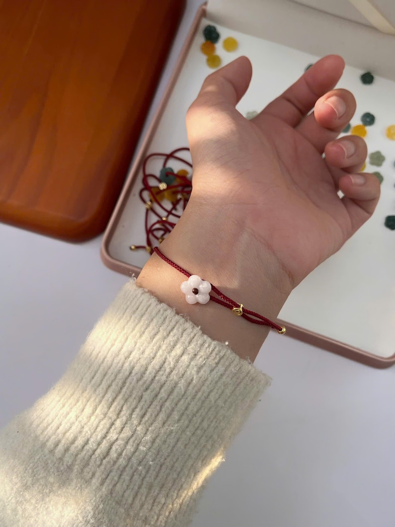 May include: A red string bracelet with a white flower charm. The bracelet features gold-colored accents and is worn on a wrist. The background includes a white surface and a tray with small, colorful decorative elements.