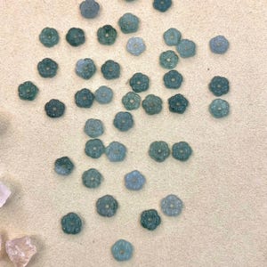 May include: A collection of small, flower-shaped beads in shades of teal and green. The beads are scattered on a light beige surface. Each bead has a detailed floral design, perfect for jewelry making or craft projects.