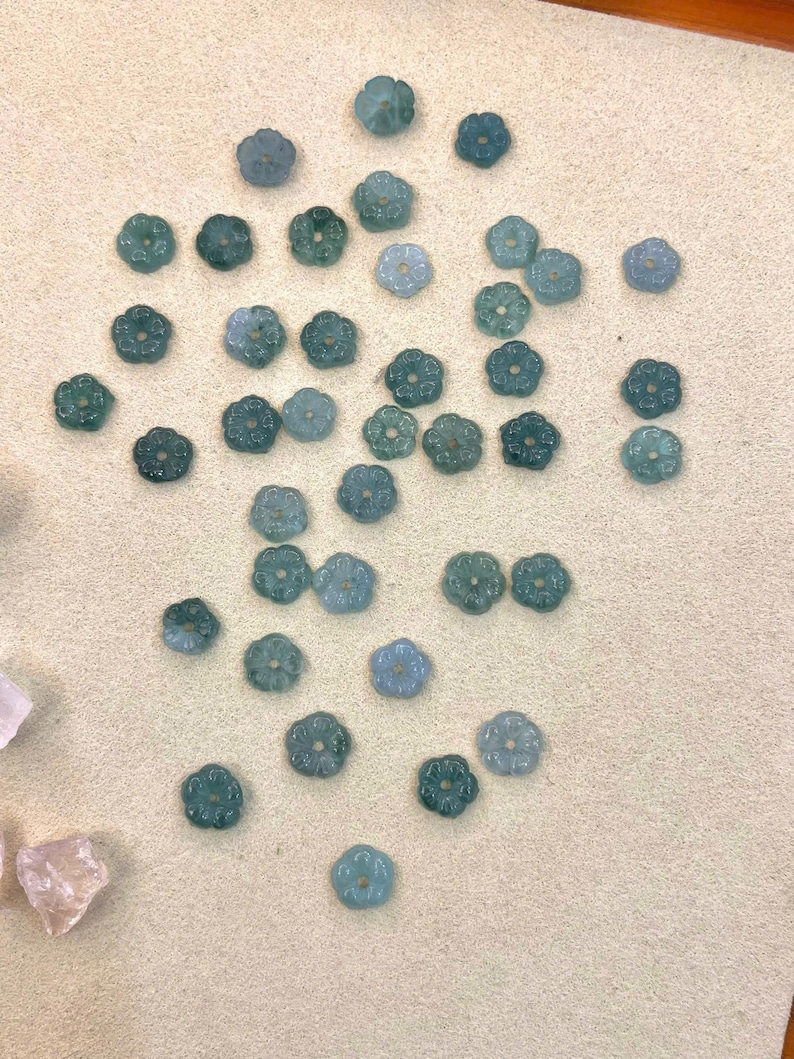 May include: A collection of small, flower-shaped beads in shades of teal and green. The beads are scattered on a light beige surface. Each bead has a detailed floral design, perfect for jewelry making or craft projects.