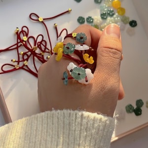 Peut inclure: Gros plan sur un bracelet en fil rouge orné de perles en forme de fleurs colorées. Les perles sont de couleurs jaune, verte et blanche. D'autres fils rouges avec des détails dorés suggèrent un thème de création de bijoux.