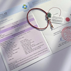May include: A red string bracelet with a carved, blue-green jade flower and gold accents. The bracelet rests on a report with text and a Vietnam Value logo. The jade flower measures 11.65 x 11.74 x 2.82 mm.