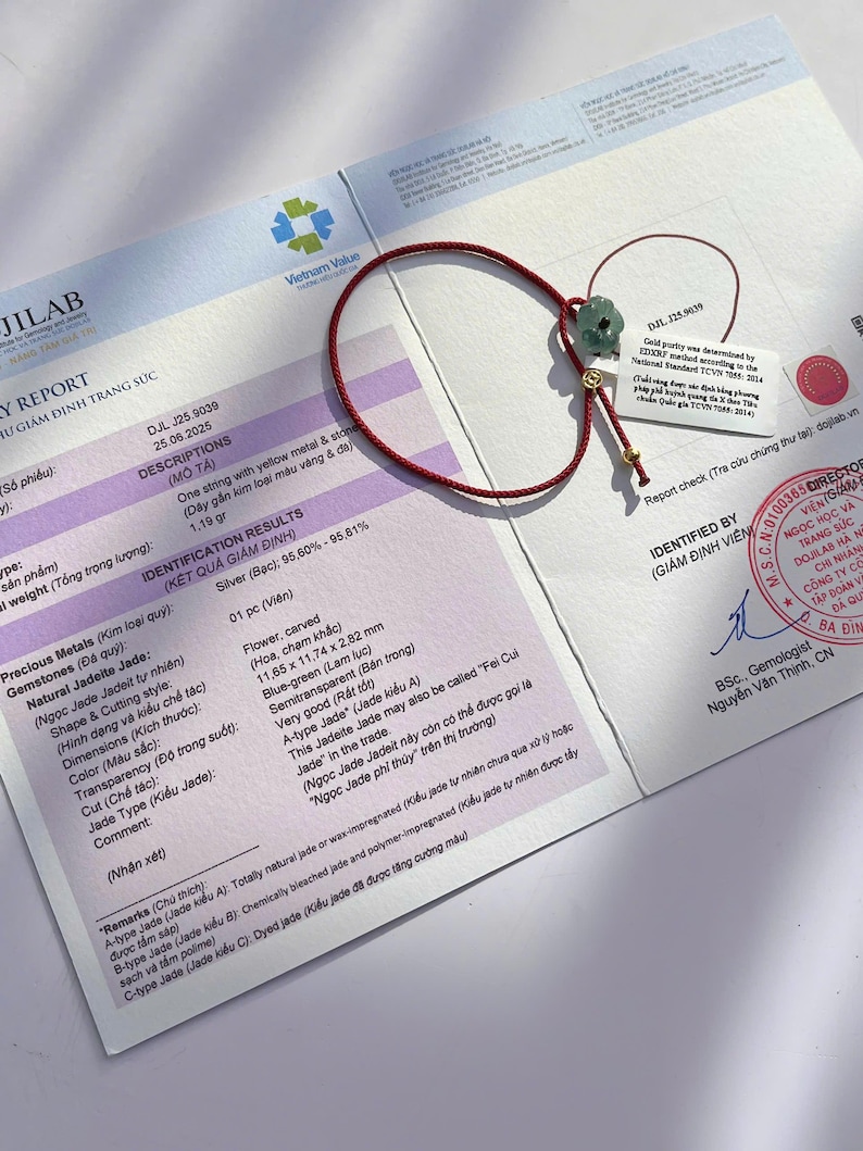 May include: A red string bracelet with a carved, blue-green jade flower and gold accents. The bracelet rests on a report with text and a Vietnam Value logo. The jade flower measures 11.65 x 11.74 x 2.82 mm.