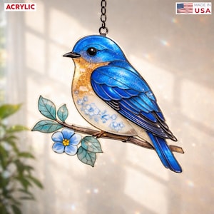 May include: An acrylic suncatcher featuring a bluebird perched on a branch with light blue flowers and green leaves. The bird has a bright blue head and wings with a gold-flecked chest and white belly. Text reads ACRYLIC and MADE IN USA.