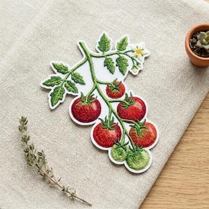 May include: An embroidered patch featuring a vine of red tomatoes with green leaves and a small yellow flower. The design shows several round red tomatoes, one partially green tomato, and one small green tomato, all connected by green embroidered stems.
