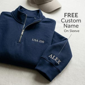 May include: Navy blue quarter-zip sweatshirt with the text "USA 250" embroidered on the chest. The sleeve features the embroidered name "ALEX". A tan baseball cap is in the background. The text "FREE Custom Name On Sleeve" is also visible.