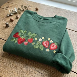 May include: A forest green crewneck sweatshirt featuring detailed embroidery of red strawberries, green leaves, and small white flowers with yellow centers. The embroidery is positioned across the chest area of the garment.