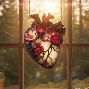 May include: A stained glass heart-shaped suncatcher with red and white floral accents. The heart features a detailed anatomical design with veins and is suspended by a chain. The suncatcher is hanging in front of a window with a blurred outdoor background.
