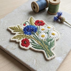 May include: Embroidered floral patch featuring a bouquet of red poppies, blue cornflowers, and white daisies with yellow centers. The patch has a cream-colored background and green stems. Two spools of thread are in the background.