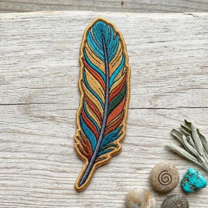 May include: Embroidered feather patch with a gold border. The feather features a vibrant design with teal, orange, yellow, and green colors. The patch is placed on a light wood surface.