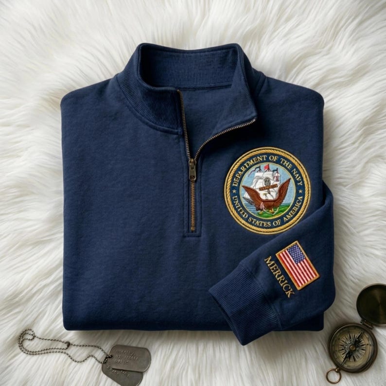 May include: Navy blue quarter-zip sweatshirt with a gold zipper. Features a Department of the Navy patch on the chest and an American flag patch on the sleeve. The sleeve also has the name "MERRICK" embroidered in gold.